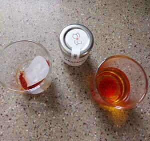 Two methods of preparing saffron. Two clear glass one containing an ice cube with crushed saffron sprinkled on the ice. The ice will melt slowly and allow the saffron to release colour and flavour slowly. The second glass contains lukewarm water with crushed saffron steeped in it. This methos will take 10 to 15 minutes for the saffron to be ready for use.